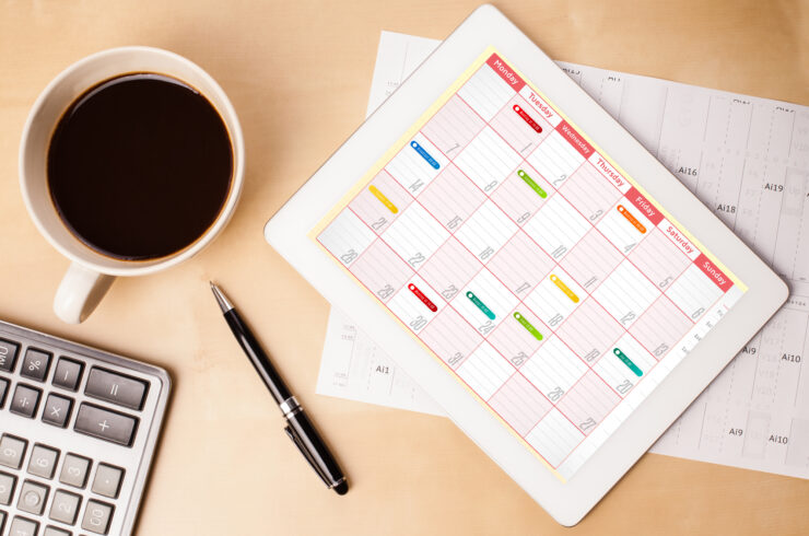 Overhead view of a desk covered with a keyword, mug of coffee, a pen, a paper calendar, and a smart tablet displaying a work schedule.