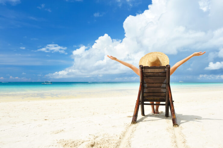 A person sitting on a wooden chair while wearing a straw sun hat on a white sand beach, clouds in the bright blue sky and the ocean visible on the horizon.