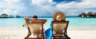 Two people are relaxing in beach chairs on a beach. They are looking out at the sea, clouds, and small vacation homes.